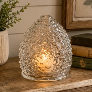 May include: A clear, dome-shaped glass lamp with a textured, bubble-like surface. The lamp features a rounded top and a circular base, emitting a warm, soft glow from an internal light source that illuminates the intricate, bumpy glass pattern.