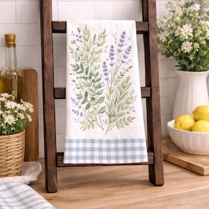 May include: A white kitchen towel with a floral design of green leaves and purple flowers, displayed on a small wooden ladder. The bottom of the towel has a blue and white checkered pattern. The ladder is dark brown and sits on a wooden countertop.