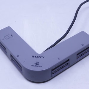 Puede incluir: Un adaptador de tarjeta de memoria Sony PlayStation gris con un cable negro. El adaptador tiene cuatro ranuras etiquetadas A, B, C y D. El logotipo de PlayStation es visible en el dispositivo.