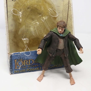 Puede incluir: Una figura de acción de plástico de un personaje de El Señor de los Anillos: El Retorno del Rey. La figura lleva una túnica marrón, una capa verde y tiene los pies descalzos. La figura está en posición de pie con los brazos extendidos. La figura está empaquetada en una caja de cartón con un mapa de la Tierra Media en la parte posterior.
