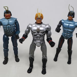 May include: Three action figures with insect-like features. The figures are in shades of blue, black, and silver, with articulated joints and insect-like antennae. The central figure is silver, flanked by two blue figures.