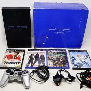 May include: A black PlayStation 2 console with a black controller, a power adapter, and four video games. The games are Rugby, Charlie's Angels, SOCOM II, and Salt Lake 2002. The console is in its original box.
