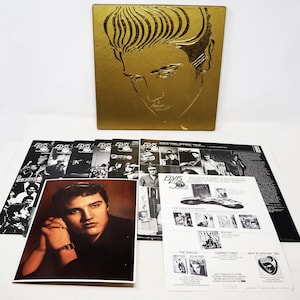Peut inclure: Une boîte dorée avec une image en noir et blanc d'Elvis Presley sur le devant. La boîte est ouverte et contient un ensemble de disques vinyles, un livret et une photo d'Elvis Presley. Les disques sont noirs et ont une étiquette dorée avec le texte "Elvis 50th Anniversary" dessus. Le livret est blanc et a une image en noir et blanc d'Elvis Presley sur le devant. La photo est en noir et blanc et montre Elvis Presley assis sur une chaise. Le texte sur le livret indique "Elvis 50th Anniversary: The Singles, Compact Discs, New in January 1995".