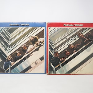 May include: Two vintage vinyl record albums. One album cover is blue and features the text "The Beatles 1967-1970". The other album cover is red and features the text "The Beatles 1962-1966". Both albums feature a photo of the band.
