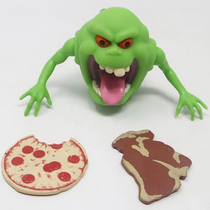 Może przedstawiać: Zielona figurka ducha z dużymi ustami i językiem, trzymająca pizzę i stek. Duch pochodzi z filmu Ghostbusters.
