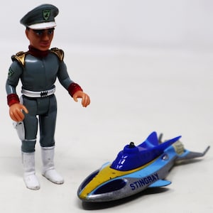 Puede incluir: Una figura de acción gris y roja de un hombre con uniforme militar y un sombrero blanco, de pie junto a un submarino de juguete azul, amarillo y plateado con la palabra "STINGRAY" impresa en el lateral.