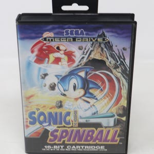 May include: A black Sega Mega Drive game cartridge box featuring Sonic the Hedgehog in a pinball-themed game. The box has a blue, red, and yellow color scheme with the game title "Sonic Spinball" prominently displayed. The box also includes the Sega logo.