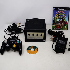 May include: A black Nintendo GameCube console with a black controller, a black power adapter, and two game discs. One disc is for the game "Super Monkey Ball" and the other is for "Double Dash!!".