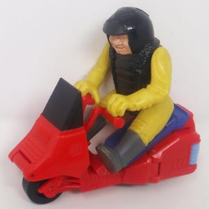 May include: A vintage toy motorcycle in red, black, yellow, and blue. The rider wears a black helmet and vest over a yellow jumpsuit. The motorcycle has a red body, black windshield, and blue seat.
