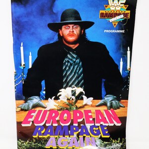 May include: A black and white program for the WWF European Rampage Again tour. The cover features a man in a black suit and hat, with a striped tie, sitting at a table with white flowers. The text "European Rampage Again" is in pink, yellow, and blue letters.