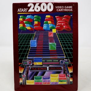 Puede incluir: Una caja de cartucho de videojuego Atari 2600 roja con una imagen colorida del juego KLAX en la parte delantera. El título del juego está en letras grandes y en negrita y la caja presenta un patrón de cuadrícula y un bono de fichas de 1.000.000.