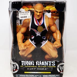 Puede incluir: Una figura de acción de Kurt Angle de 35,5 cm con un atuendo de lucha libre. La figura lleva un maillot de lucha libre negro y azul con estrellas y rayas. La figura lleva una medalla de oro alrededor del cuello. La figura está en una postura de lucha libre. El embalaje dice "Ring Giants" y "13 puntos de articulación".
