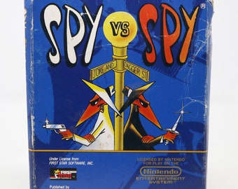 Può includere: Una scatola di cartuccia di gioco blu e bianca per il sistema di intrattenimento Nintendo. La scatola presenta il titolo "SPY VS SPY" in grandi lettere bianche. La parte anteriore della scatola mostra due spie dei cartoni animati, uno in bianco e uno in nero, che si fronteggiano su un angolo di strada. Il testo "Under License from FIRST STAR SOFTWARE, INC." e "LICENSED BY NINTENDO FOR PLAY ON THE Nintendo ENTERTAINMENT SYSTEM" è stampato sulla scatola.