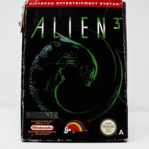 May include: A black box with a green alien creature on the front. The box is for a Nintendo Entertainment System game called "Alien 3".