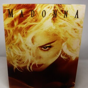 Puede incluir: Un primer plano de una portada de álbum vintage de Madonna con una foto de Madonna con cabello rubio y una expresión seria. El título del álbum "MADONNA" está impreso en letras doradas en la parte superior de la portada.