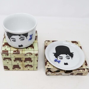 Puede incluir: Un juego de taza y platillo de porcelana blanca con una imagen en blanco y negro de Charlie Chaplin. La taza y el platillo están en su caja original, que presenta un patrón repetido de la cara de Chaplin.