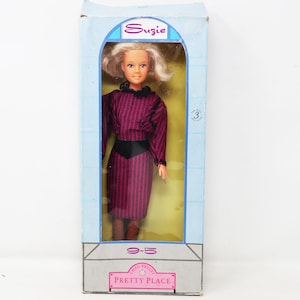 May include: A Suzie doll in a light blue box with the text Pretty Place and 9-5. The doll has blonde hair and wears a long-sleeved purple dress with a black pattern, a black waist belt, and a black collar. The box indicates for ages 3 and over.