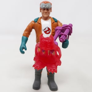 Puede incluir: Figura de acción de los Cazafantasmas con un personaje con un mono naranja, guantes turquesa y botas grises. La figura sostiene un paquete de protones morado y un fantasma rojo. La camiseta del personaje tiene el logo de los Cazafantasmas.
