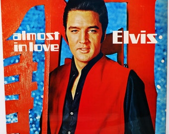 Può includere: Copertina dell'album "Almost in Love" di Elvis. La copertina presenta un ritratto di Elvis Presley con un gilet rosso e una camicia nera su uno sfondo rosso e blu. Il titolo dell'album e il nome dell'artista sono ben visibili.