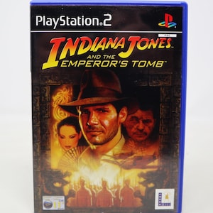 May include: A PlayStation 2 video game case featuring Indiana Jones and the Emperor's Tomb. The cover art shows Indiana Jones in a fedora, with a woman and a man behind him. The game is rated 11+.