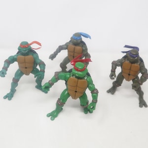Op de afbeelding: Vier Teenage Mutant Ninja Turtles actiefiguren. Elke figuur is groen met een bruin schild en een gekleurd masker: rood, blauw, paars en groen. De figuren staan in dynamische poses, klaar voor actie.