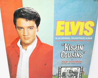 Può includere: Una copertina di un album di vinile vintage con Elvis Presley che indossa una giacca rossa. Il titolo dell'album è "Kissin' Cousins" ed è una colonna sonora del film Metro-Goldwyn-Mayer. La copertina dell'album è di colore blu chiaro e verde.