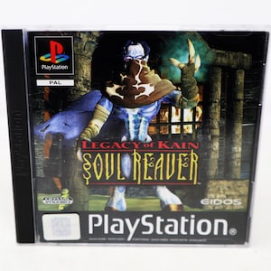 Może przedstawiać: Pudełko z grą PlayStation "Legacy of Kain: Soul Reaver". Okładka przedstawia postać z niebieską skórą z brązowo-białą maską na tle mrocznego, gotyckiego tła. Widoczne jest logo PlayStation.
