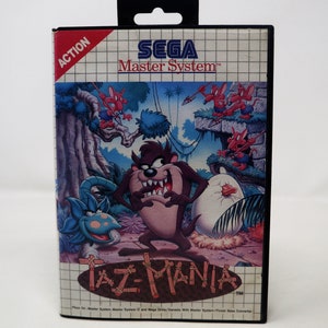 Pode incluir: Um cartucho de jogo Sega Master System com Taz-Mania. A parte frontal do cartucho apresenta uma ilustração colorida de Taz, o Diabo da Tasmânia, com um grupo de pequenas criaturas vermelhas semelhantes a pássaros no fundo. O texto "ACTION" está no canto superior esquerdo, e "SEGA Master System™" está no canto superior direito. O título do jogo "Taz-Mania™" está na parte inferior central do cartucho.