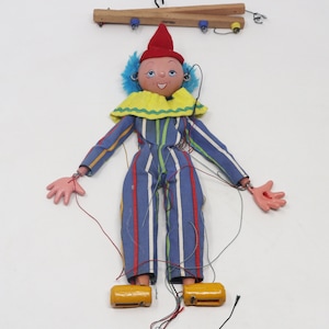 Puede incluir: Un títere a rayas azules y blancas con un sombrero rojo, un cuello amarillo y pelo azul. El títere tiene una cara pintada con una sonrisa y está unido a cuerdas para su manipulación.