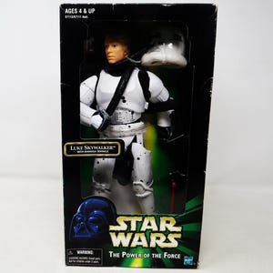 Op de afbeelding: Een Star Wars actiefiguur van Luke Skywalker in een wit en zwart Stormtrooper-outfit, verpakt in een zwarte doos. De doos heeft het Star Wars-logo en de tekst "The Power of the Force". De figuur bevat een afneembare helm.