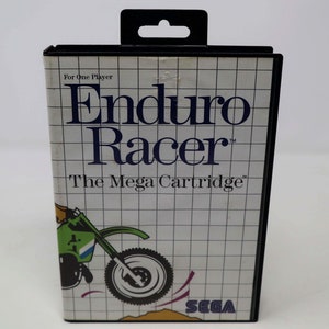 May include: A black Sega Genesis game cartridge with a white label. The label features a cartoon illustration of a person riding a dirt bike. The text on the label reads "Enduro Racer", "The Mega Cartridge", and "Sega".