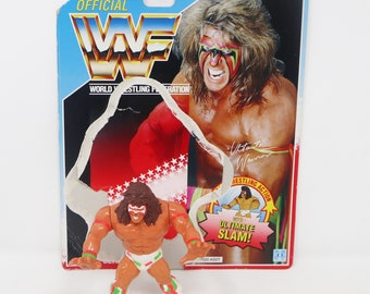 May include: A vintage action figure of Ultimate Warrior from the World Wrestling Federation (WWF). The figure is wearing a red, white, and green wrestling outfit with a red and yellow cape. The figure is posed in a fighting stance with its arms raised. The figure is packaged in a cardboard box with a picture of Ultimate Warrior on the front. The box also includes the text "OFFICIAL WWF" and "WORLD WRESTLING FEDERATION".