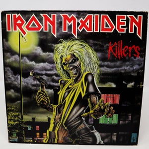 May include: A vintage vinyl record album cover for the band Iron Maiden. The cover features a yellow and black illustration of Eddie, the band's mascot, holding a bloody hammer. The album title "Killers" is written in red letters on the cover.