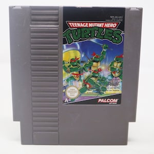 May include: A Teenage Mutant Hero Turtles video game cartridge for the Nintendo Entertainment System. The cartridge is gray with a black label featuring a colorful image of the four turtles in action. The label also includes the game title, the Nintendo logo, and the PALCOM software logo.