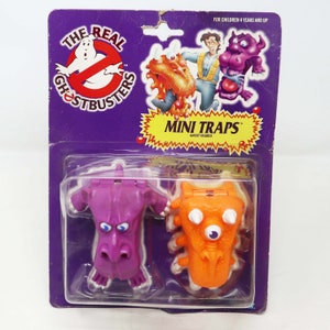 Peut inclure: Un emballage en plastique contenant deux petites figurines de fantômes Ghostbusters Mini Traps, l'une violette et l'autre orange. L'emballage a un fond violet avec les mots "The Real Ghostbusters" et "Mini Traps Ghost Figures" imprimés dessus.