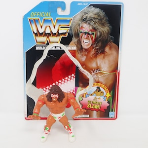 May include: A vintage action figure of Ultimate Warrior from the World Wrestling Federation (WWF). The figure is wearing a red, white and green wrestling outfit with a red and yellow cape. The figure is posed in a fighting stance with its arms raised. The figure is packaged in a cardboard box with a picture of Ultimate Warrior on the front. The box also includes the text "OFFICIAL WWF" and "WORLD WRESTLING FEDERATION".