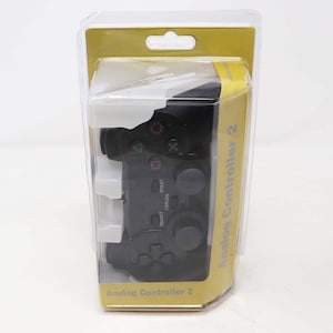 May include: A black PlayStation 2 analogue controller in a clear plastic package with a yellow background. The package says "Analogue Controller 2".