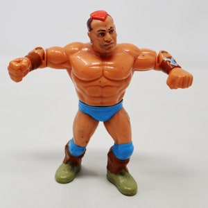 May include: A plastic action figure of a wrestler with a red mohawk, brown wristbands, blue trunks, and blue knee pads. The figure has a muscular build and is posed in a fighting stance.