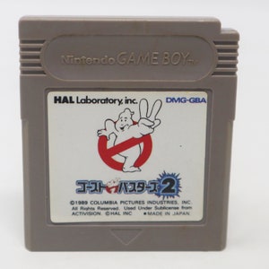 Puede incluir: Un cartucho gris de Game Boy Color con una etiqueta blanca. La etiqueta presenta un fantasma de dibujos animados con un círculo rojo y una barra diagonal. El texto de la etiqueta dice "HAL Laboratory, inc." "DMG-GBA" "ゴーストバスターズ2" "©1989 COLUMBIA PICTURES INDUSTRIES, INC." "All Rights Reserved. Used Under Sublicense from ACTIVISION. ©HAL INC. MADE IN JAPAN."