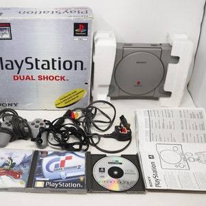 May include: A grey PlayStation console with a DualShock controller, two game discs, and a power cable. The console is in its original box, which is labelled "PlayStation DualShock".