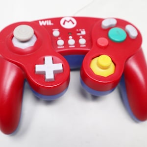 Puede incluir: Mando de videojuegos rojo y azul con una cruceta direccional blanca y un joystick gris. El mando tiene un joystick amarillo y varios botones. La palabra "WIL" y un logotipo "M" son visibles en el mando.