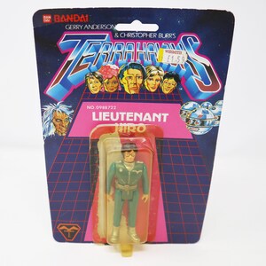 Puede incluir: Una figura de acción de plástico del teniente Hiro de la serie de televisión Terrahawks. La figura lleva un uniforme verde con un cinturón negro y un casco plateado. La figura está empaquetada en una tarjeta con un fondo rosa y una imagen de los personajes de Terrahawks.