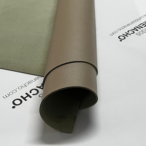 Puede incluir: Una lámina enrollada de material color taupe con una superficie texturizada. El borde revela un reverso verde oscuro. El material está sobre una superficie blanca con el texto "MENACHO" y "www.cuidadosmenacho.com" impreso.