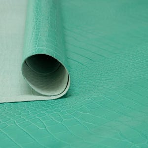 Puede incluir: Un rollo de tela de cuero sintético con estampado de cocodrilo turquesa. La tela tiene una superficie texturizada que se asemeja a la piel de cocodrilo.
