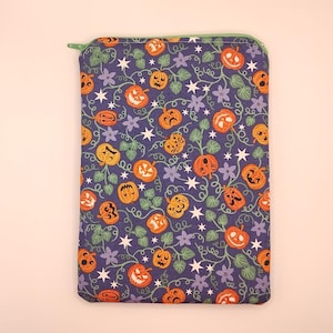 May include: A rectangular fabric pouch featuring a dark purple background with a repeating pattern of orange jack-o'-lanterns, green vines, small light purple flowers, and white four-pointed stars. The pouch has a light green zipper at the top.