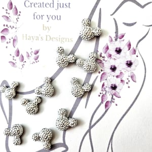 Può includere: Pesi per velo color argento a forma di Mickey Mouse, sparsi su uno sfondo bianco con accenti floreali e testuali. Il testo recita "Veil Weights, Created just for you by Haya's Designs."