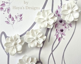 May include: White floral veil weights. The image shows five white, flower-shaped veil weights. The text at the top reads "Veil Weights" and the text below reads "Created just for you by Haya's Designs". The background has a floral design.