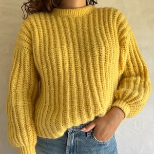 Puede incluir: Un suéter de punto amarillo con textura de canalé y mangas largas. El suéter tiene un cuello redondo y un ajuste holgado.