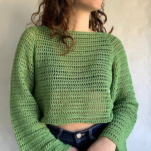 May include: A green crochet top with long sleeves and a loose fit. The top is made with a delicate open stitch pattern.