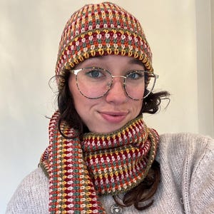 May include: A handmade crochet hat and scarf set. The hat and scarf feature a colorful pattern with red, yellow, green, and orange hues. The scarf is wrapped around the neck. The hat is on the head.
